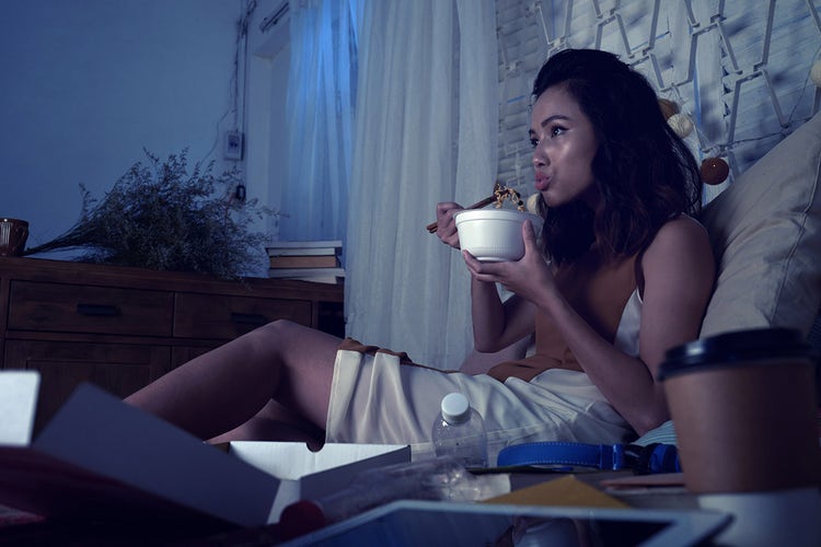 Person sitting on a bed and eating takeout food in a dimly lit room