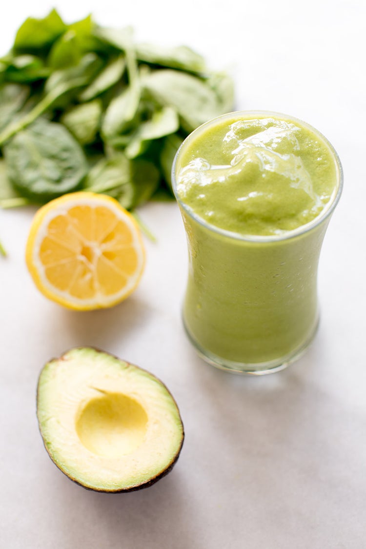 A table with a green ginger smoothie in a glass, a sliced avocado, a sliced orange, and a bunch of spinach
