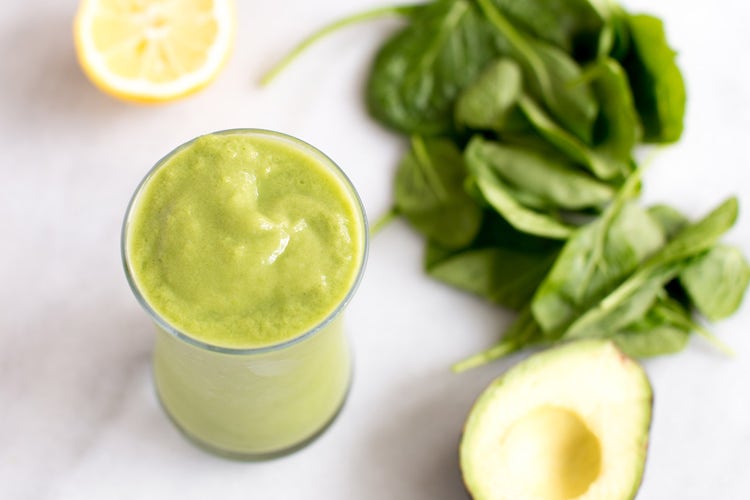 A table with a green ginger smoothie in a glass, a sliced avocado, a sliced orange, and a bunch of spinach