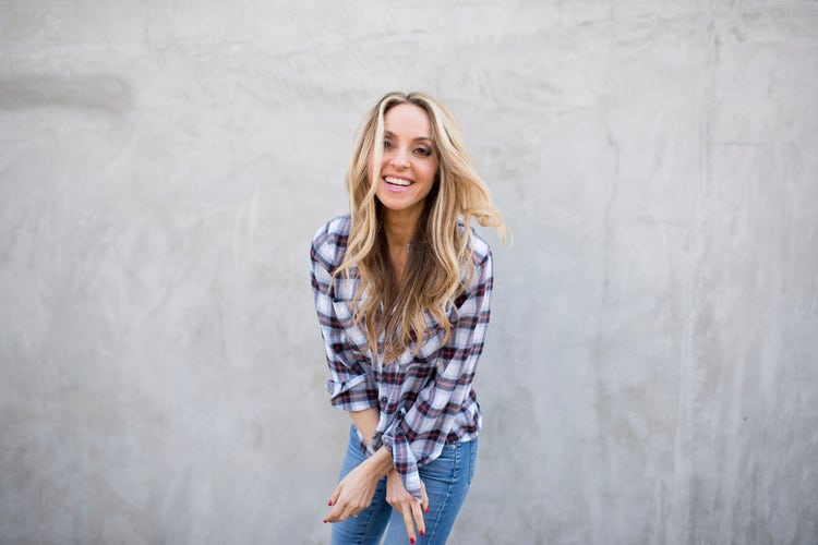 Gabrielle Bernstein smiling and wearing a blue plain shirt and standing against a grey wall