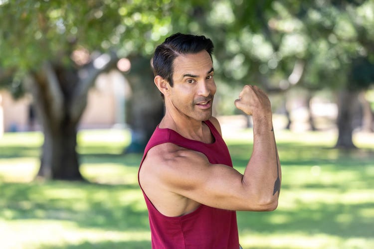 Jorge Cruise flexes his right bicep while standing in a park. Red tank top, short black hair